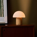 Cordless table lamp Ronald with light brown wood base and frosted glass dome shade glowing on a wooden cabinet corner beside a stack of books and framed artwork