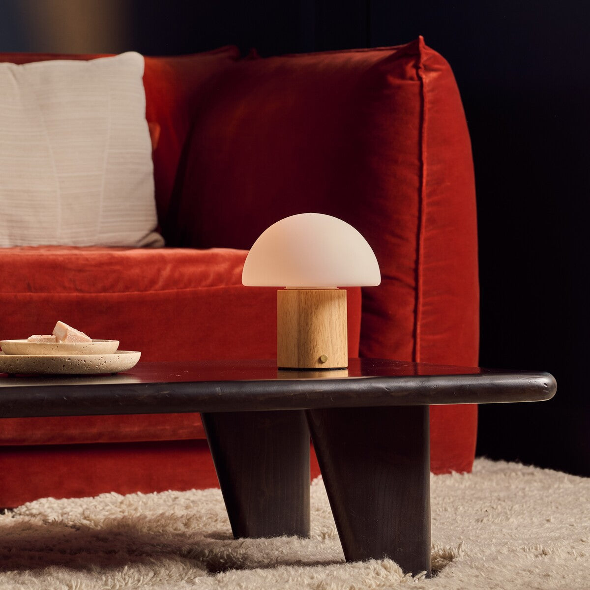 Rechargeable table lamp Ronald with light brown wood base and frosted glass dome shade on a dark coffee table beside a red velvet sofa