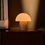 Rechargeable table lamp Ronald with light brown wood base and frosted glass dome shade glowing softly on a wooden sideboard beside a framed picture