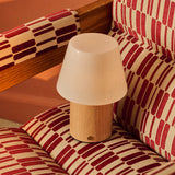 Cordless table lamp Jadey Medium, light wood base with opal glass shade on red patterned armchair