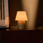 Cordless table lamp Jadey Medium, light wood base and glass shade glowing on dresser near blinds