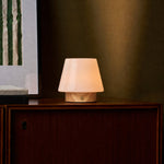 Oplaadbare tafellamp Jadey Small, licht hout met glazen kap op donkere kast bij ingelijste kunst