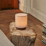 Oplaadbare tafellamp Dick Small Dark Brown, linnen kap op donker houten voet, op rustieke boomstam bijzettafel