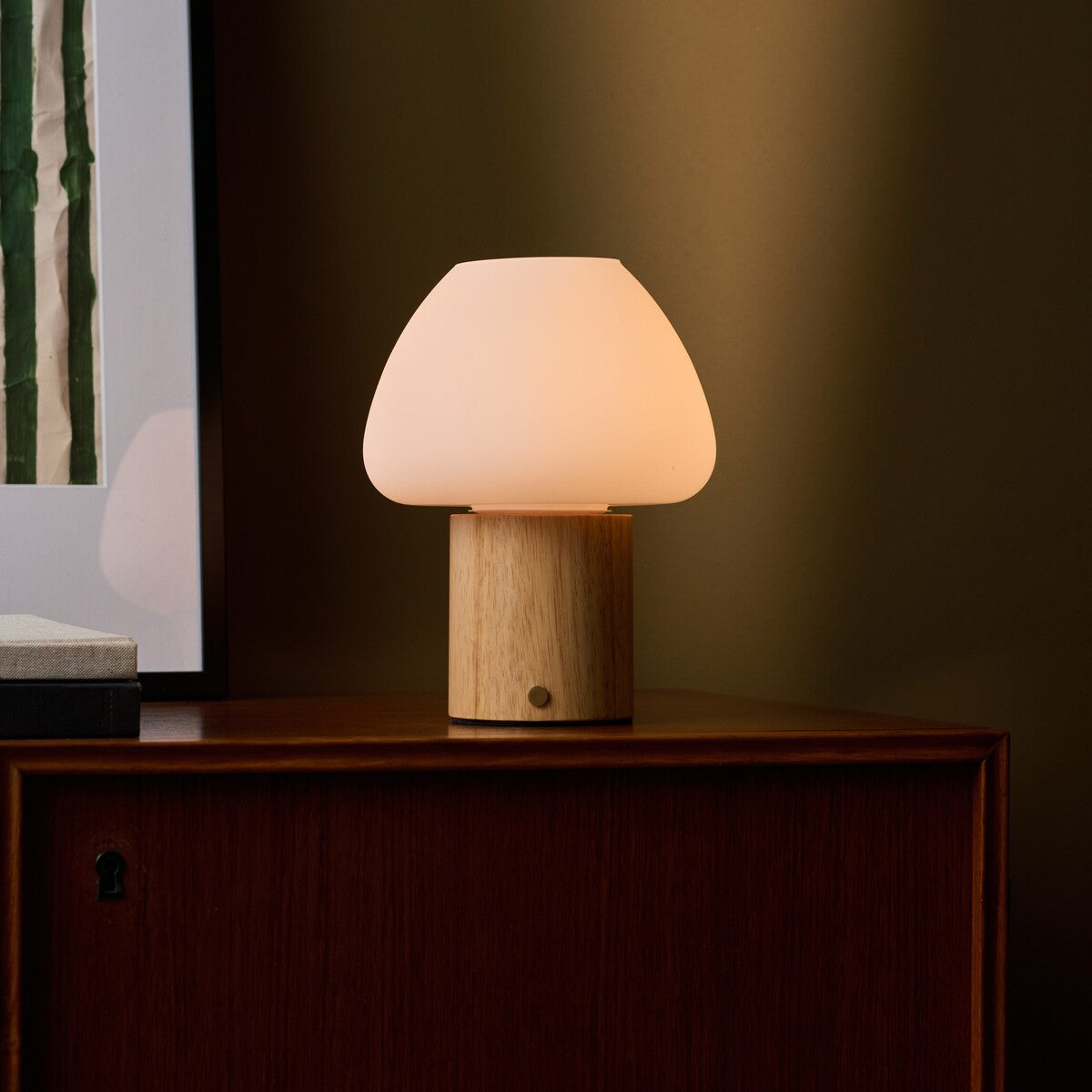 Fynn cordless table lamp glowing warm light, light brown wooden base beside framed wall art