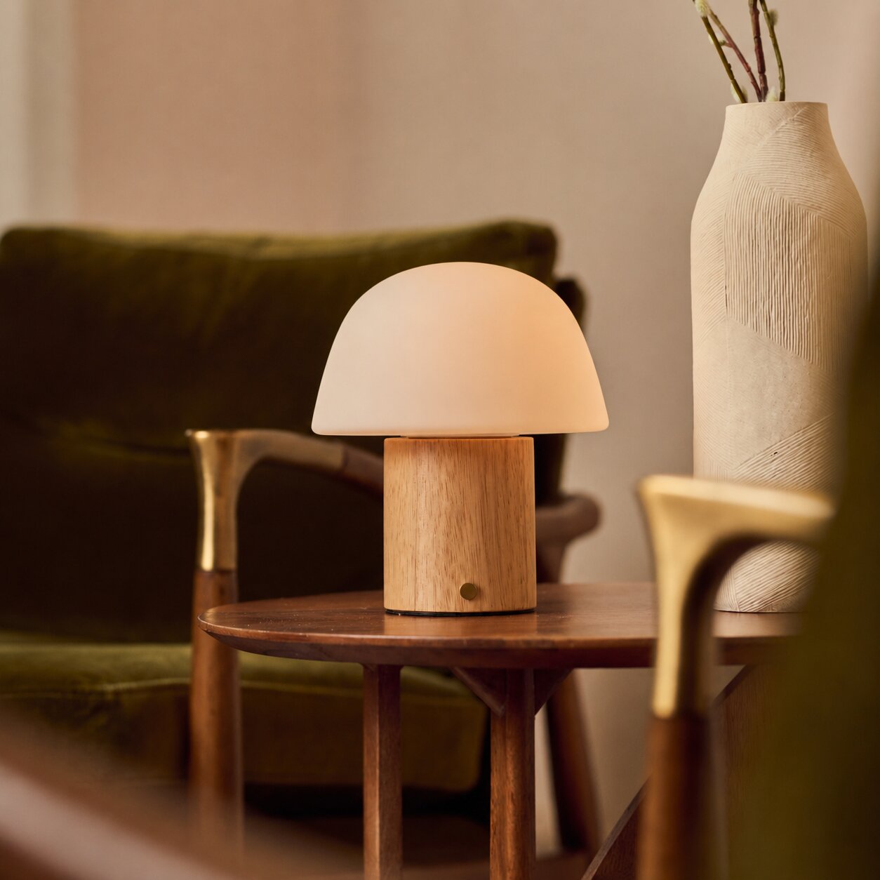 Rechargeable table lamp Ton with light brown wood base and frosted glass dome shade on a wooden side table beside a green velvet sofa and a ceramic vase