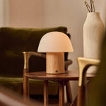 Rechargeable table lamp Ton with light brown wood base and frosted glass dome shade on a wooden side table beside a green velvet sofa and a ceramic vase