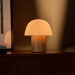 Cordless table lamp Ton with light brown wood base and frosted glass dome shade glowing softly on a dark wooden sideboard beside a framed picture