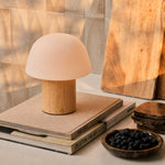 Rechargeable table lamp Ton with light brown wood base and frosted glass dome shade glowing on a stack of books beside a wooden cutting board and a bowl of blueberries