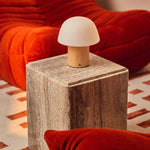 Cordless table lamp Ton with light brown wood base and frosted glass dome shade on a travertine side table beside a burnt-orange curved sofa