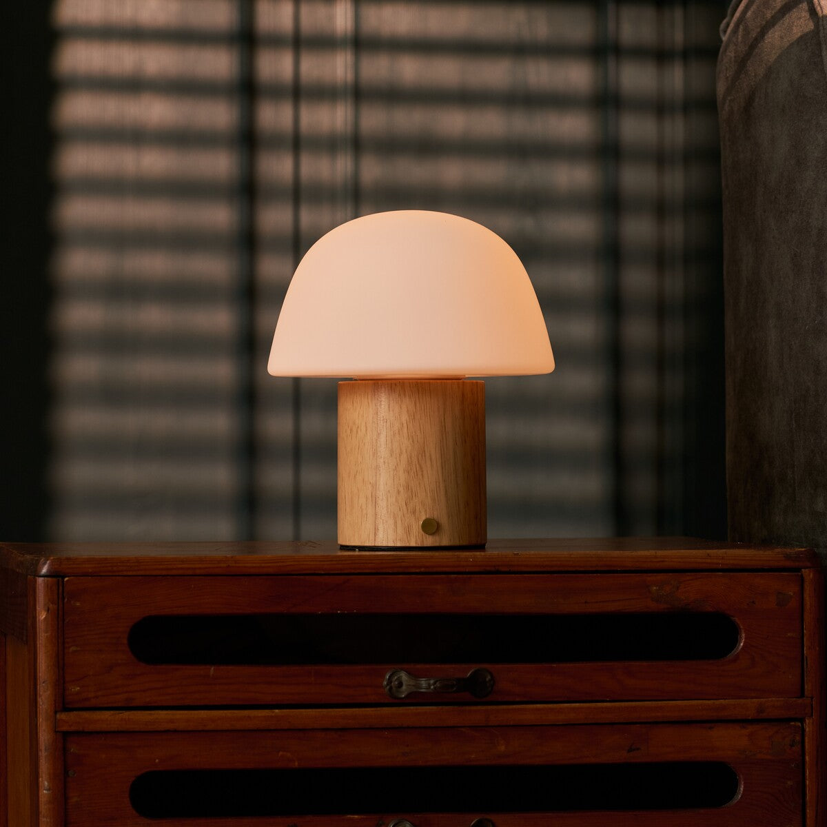 Rechargeable table lamp Ton with light brown wood base and frosted glass dome shade glowing on a wooden dresser with slatted light and shadow behind