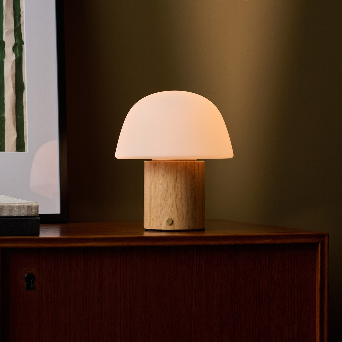 Rechargeable table lamp Ton with light brown wood base and frosted glass dome shade glowing on a wooden sideboard against an olive green wall beside a framed picture