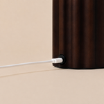 Gros plan sur le socle en bois cannelé brun foncé d'une lampe Tavellio – détail du connecteur de charge USB-C et de son câble blanc